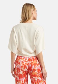Cremfarbener, gekürzter Pullover mit Fledermausärmeln, der eine Mittelnaht auf der Rückseite hat. Kombiniert mit floral gemusterten Hosen in Orange und Pink.
