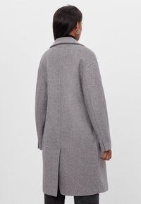 Šedý vlněný kabát se střihem do rovna, klopovým límcem, dlouhými rukávy a rozparkem na zádech. Textura působí měkce s broušeným povrchem.