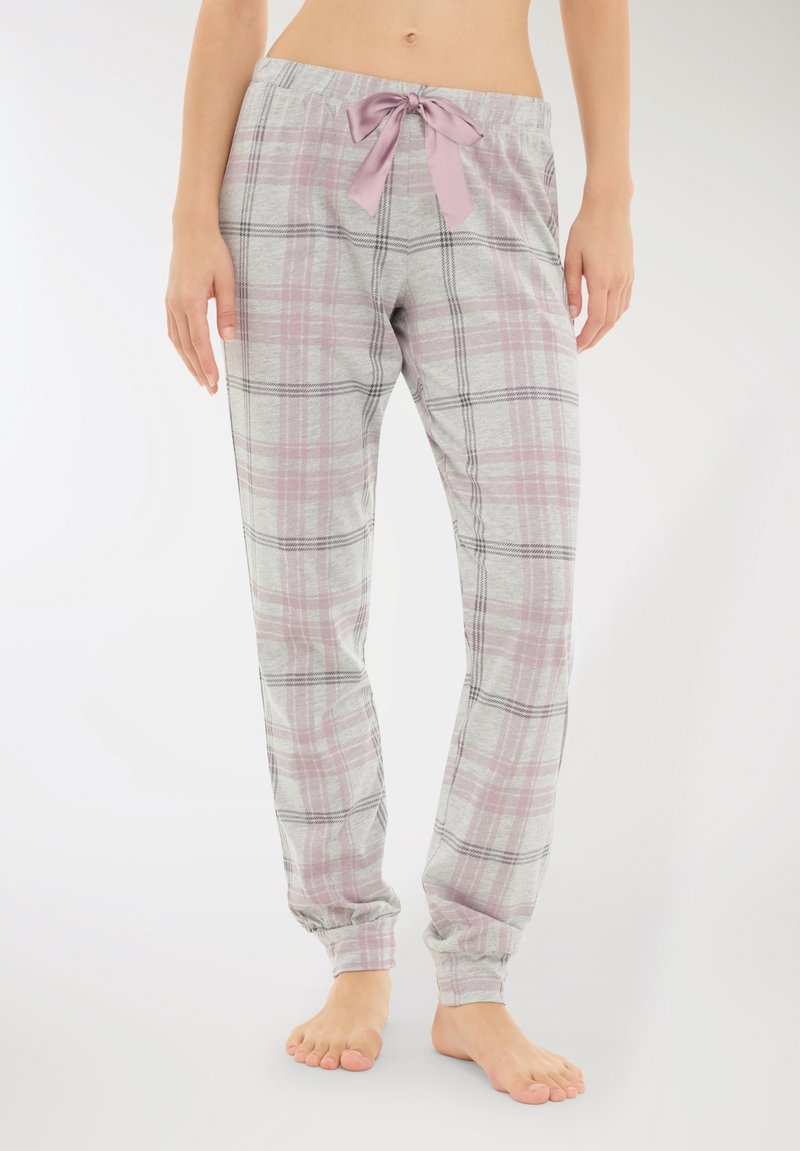 Donna che indossa pantaloni da pigiama a quadri grigi e rosa con un fiocco di raso in vita, in piedi a piedi nudi su uno sfondo bianco.