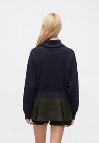 Tumši zils džemperis ar augstu apkakli, garām piedurknēm un īsu piegriezumu, kopā ar teksturētu, krokojumu tumšu svārku.