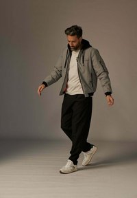 Veste bomber grise avec capuche, accents noirs, t-shirt blanc, pantalon noir et baskets blanches, dans un cadre de studio neutre.