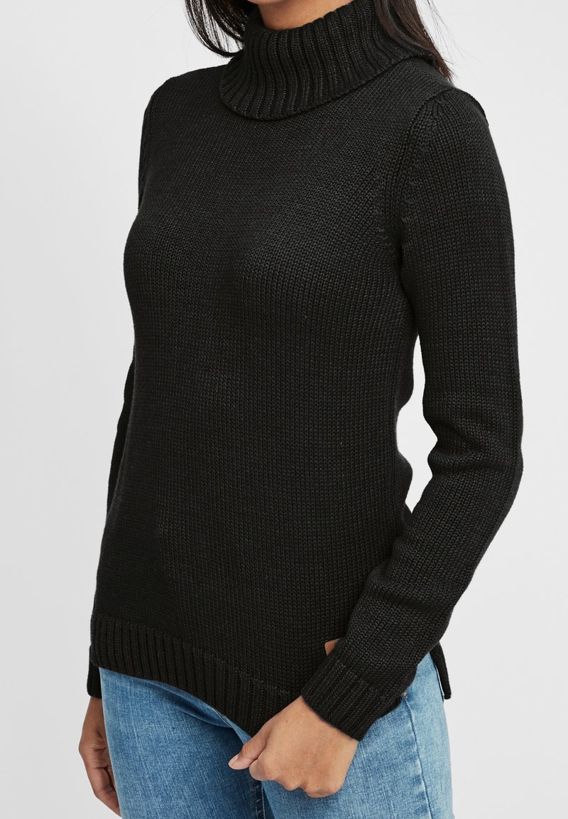 Femme portant un pull col roulé noir et un jean bleu, se tenant devant un fond clair.