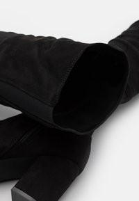 Botas negras de ante hasta la rodilla con un tacón robusto. Presentan un interior suave y paneles elásticos para un mejor ajuste. Detalle de cremallera en el costado.