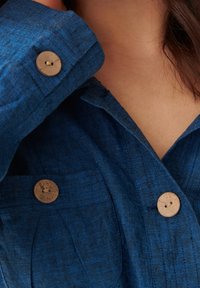 Blå linne skjorta med två korkknappar framtill, med ett texturerat tyg och en krage. Närbild på övre bröstområdet.
