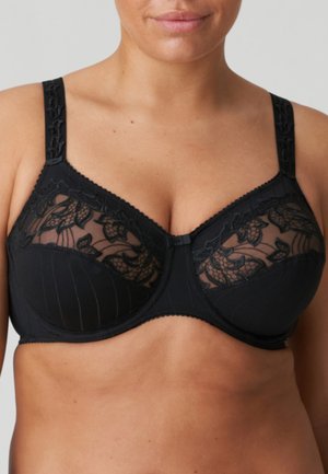 Vrouw draagt een zwarte beugelbh met bloemenkantmesh op de bovenste cups en verstelbare brede bandjes tegen een effen achtergrond.