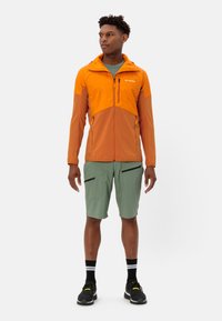 Giacca leggera arancione e marrone con zip completa e tasche, abbinata a pantaloni corti verdi con dettagli in zip neri e cintura elastica.