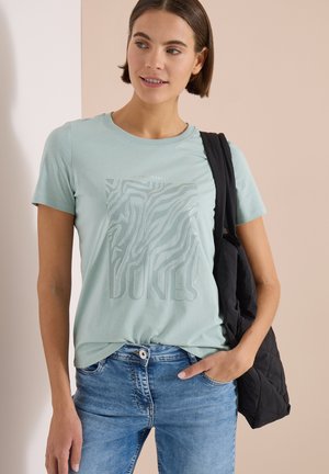 Femme portant un t-shirt graphique vert clair, un jean bleu et un sac à bandoulière noir matelassé, debout contre un mur beige et blanc.