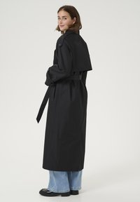 Cappotto nero a trench con vita a cintura, sezione superiore corta, dettagli a bottoni sulle maniche e texture liscia. Indossato sopra jeans a gamba larga blu.