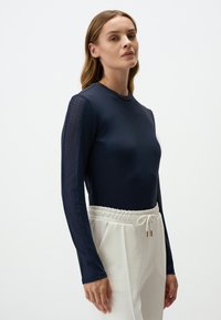Marinefarbener Langarmtop aus glattem Material mit Netzdetails an den Ärmeln, kombiniert mit off-white Hosen mit Kordelzug.