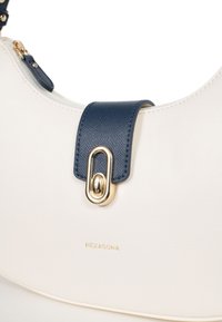 Borsa bianca con finitura testurizzata, dotata di una tracolla blu scuro e hardware dorato. Logo "HEXAGONA" impresso in oro sul davanti.
