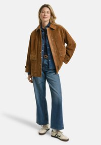 Braune Wildlederjacke mit Kragen, oversized Schnitt, zwei Taschen vorne, kombiniert mit einem Denimhemd und weiten Jeans sowie cremfarbenen Sneakers.