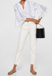 Blouse en lin blanc à rayures bleues avec détails en œillets, associée à un jean taille haute beige clair et un sac seau noir. Sandales plates.