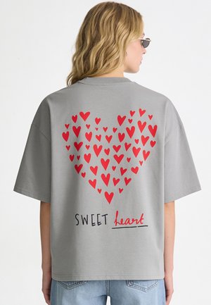 Blond meisje draagt een grijs T-shirt met een rood hart van kleine hartjes en de tekst "SWEET heart" op de achterkant, gecombineerd met blauwe spijkerbroek.