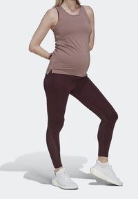 Těhotenské tílko v barvě mauve, spárované s vysokými bordovými legínami. Modelka má na sobě bílé sportovní boty, čímž předvádí elegantní, přiléhavý siluetu.