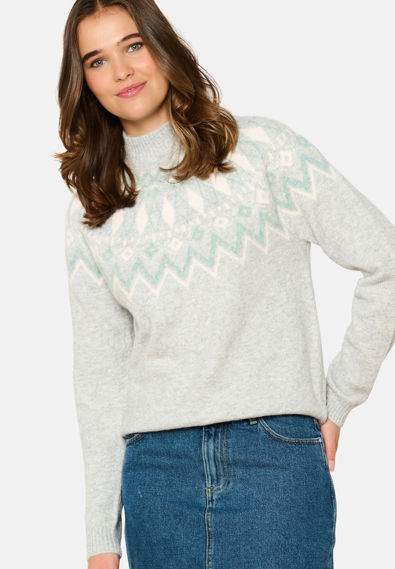 Grau gestrickter Pullover mit einem hellblauen und weißen geometrischen Muster am Kragen, mit hohem Hals und gerippten Bündchen. Über einem Jeansrock getragen.