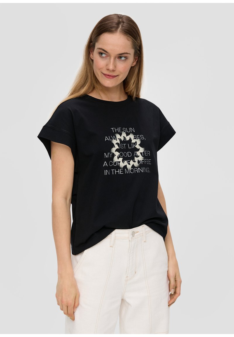Schwarzes Baumwoll-T-Shirt mit kurzen Ärmeln, mit einem weißen gedruckten Textdesign in der Mitte. Kombiniert mit hellen Hosen.