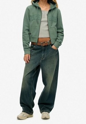 Personne portant un sweat à capuche court vert, un débardeur court gris, un jean large délavé avec une ceinture marron et des baskets blanches, debout les mains dans les poches.