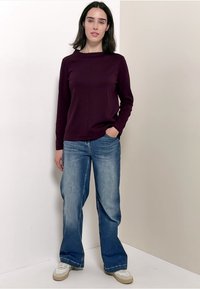 Langärmliges bordeauxfarbenes Oberteil mit hohem Kragen und lockerer Passform, kombiniert mit hellblauen, weit geschnittenen Jeans und weißen Sneakers. Schlichter Hintergrund.