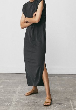 Personne debout, les bras croisés, portant une robe midi noire sans manches avec une fente latérale et des sandales plates marron sur un sol en béton.