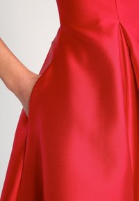 Robe en satin rouge avec un corsage ajusté et une jupe évasée. Comprend une poche sur le côté et une texture lisse avec un léger éclat.