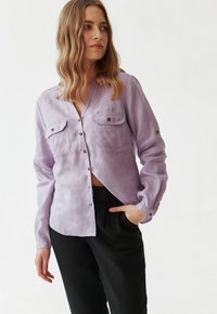 Vrouw in een lavendelkleurig overhemd met borstzakken en zwarte broek, staand met één hand in de zak, kijkend naar de zijkant.