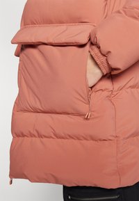 Vycpaná bunda z tlmeného ružového materiálu, s prednou vreckom na klopu a elastickými manžetami, hladká textúra so slabým leskom.