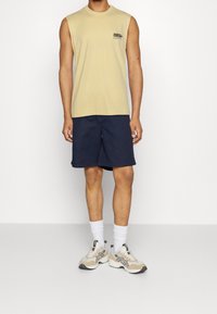 Person wearing a beige sleeveless shirt with black text, navy blue shorts, white socks, and beige and white sneakers, standing against a plain background.