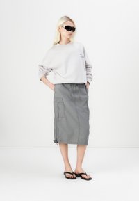 Felpa oversize grigia chiara con logo ricamato, abbinata a una gonna cargo grigia fino al ginocchio con tasche laterali e cordoncino. Infradito neri.