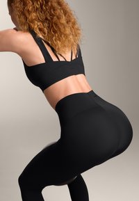 Musta urheilupaitojen yläosa, jossa on useita nauhoja, ja korkeavyötäröiset mustat legginsit. Sileä pinta, muotoon ommeltu malli, sopii liikuntaan.