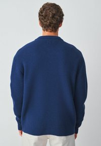 Granatowy sweter wykonany na drutach o prążkowanej teksturze, w oversize'owym kroju i szerokim kołnierzu, pokazany od tyłu na jasnym tle.