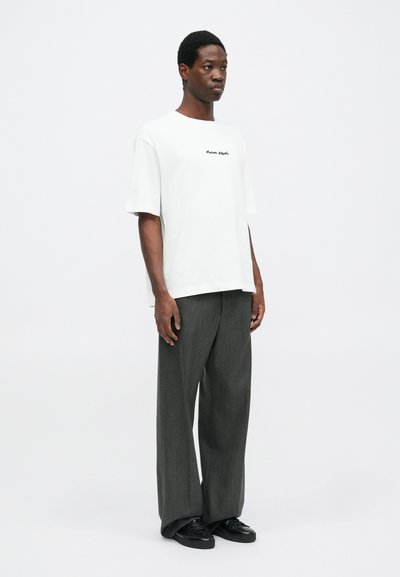 White cotton t-shirt with short sleeves, featuring a black printed logo on the chest. Paired with wide, gray pleated trousers and black shoes.