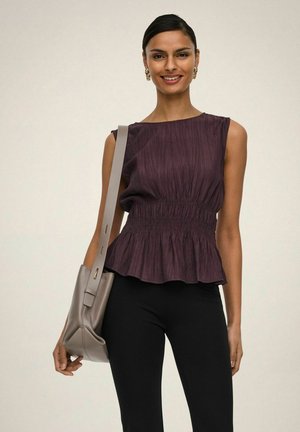 Lachende vrouw met een mouwloos paars topje en zwarte broek, met een grijze schoudertas tegen een effen achtergrond.