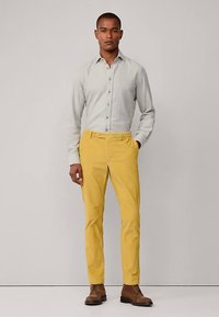 Lichtgrijs overhemd met knopen gecombineerd met gele broek en bruine schoenen. De broek heeft een strakke pasvorm en zichtbare frontzakken.