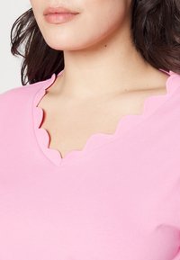 Rosa t-shirt med en scalloped V-halsdesign. Tyget är slätt och elastiskt, vilket framhäver halsringningens detalj.