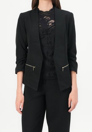 Femme portant un blazer noir avec des poches zippées dorées sur un top en dentelle noire et un pantalon noir, debout devant un fond blanc.