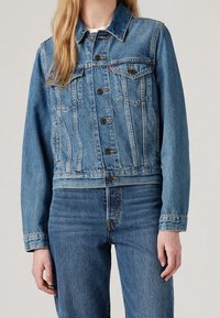Džínsová bunda světle modré barvy s klasickým střihem, knoflíkovým zapínáním, náprsními kapsami a mírným prosvětlením pro texturu. Stylizováno s džínsovými kalhotami.
