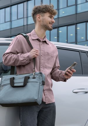 Junger Mann in pinkem Hemd, der ein Smartphone und eine blaue Umhängetasche hält, steht neben einem weißen Auto in einer städtischen Umgebung.