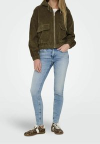 Olivgrön kordjacka med kapad längd och huva, två framfickor, i kombination med ljusblå tighta jeans och sneakers med leopardmönster.