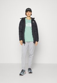 Sort pufferjakke over en mintgrøn t-shirt med hvidt logo, grå sweatsbukser og grå-blå sneakers med multicolore snørebånd. Sort kasket.