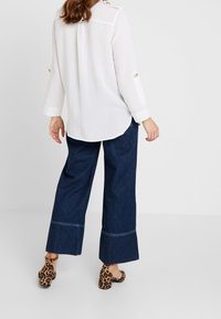 Camisa blanca de manga larga con detalles de botones, combinada con pantalones de mezclilla oscuros de pierna ancha con dobladillo acampanado y tacones de leopardo.