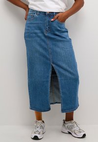 Jeansrock in mittelblau mit einem Schlitz vorne, geradem Schnitt, zwei seitlichen Taschen und abgerundetem Saum. Kombiniert mit hellgrauen Turnschuhen.
