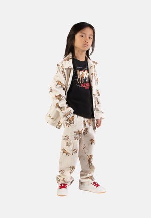 Tenue beige composée d'une veste et d'un pantalon imprimés de motifs chevaux. Chemise noire avec un design graphique et un texte rouge. Baskets blanches avec des accents rouges.