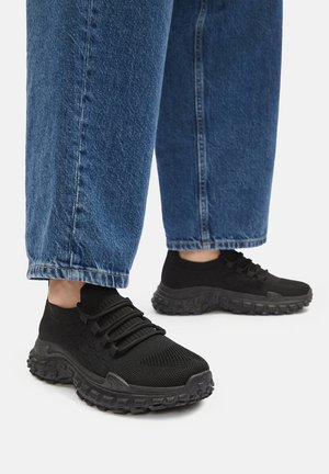 Czarne sportowe sneakersy z siateczkową cholewką, miękką teksturowaną podeszwą i elastycznymi sznurowadłami, noszone z luźnymi dżinsami.