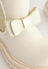 Helle Creme-Ankleboots mit texturiertem Finish. Verfügt über einen goldenen quadratischen Akzent und ein Schleifendetail an der Seite. Gummisohle mit geriffeltem Rand.