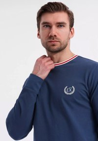 Navy long-sleeve shirt, ribbed texture, featuring a white logo with a laurel design and a red and white striped neckline.