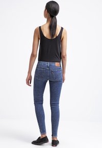 Jeans de denim azul de corte ajustado con una cintura de altura media, que cuentan con bolsillos, un parche de cuero con logo y un desgaste sutil. Combinados con una camiseta sin mangas negra.