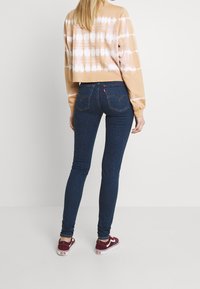 Jeans skinny azul oscuro con costuras prominentes y bolsillos traseros, combinados con un sudadera corta de teñido anudado en marrón claro y zapatillas burdeos.