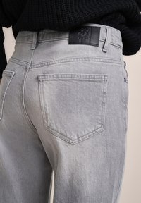 Graue Denim-Jeans im Straight-Leg-Design, mit einem Lederpatch auf der Rückseite und normalen Taschen. Der Stoff hat eine leicht strukturierte Oberfläche.