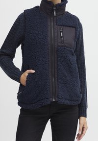 Osoba v námořnickém modrém fleecovém vestě s texturou, zapínáním na zip a kapsami, přes pletený svetr s dlouhým rukávem a černé kalhoty.