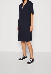 Robe bleu marine à manches courtes avec col polo et fentes sur les côtés, associée à des baskets bleu marine avec des lacets blancs et une semelle blanche.